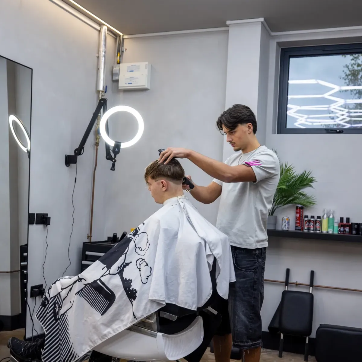 Evan clipper-fading a client's hair, product shelf and plant behind